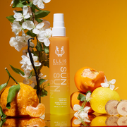 Sun Hair and Body Fragrance Mist 100ml on mirror next to oranges, lemons, cedar wood, white flowers, green leaves, and vanilla beans, on orange and yellow gradient background.
