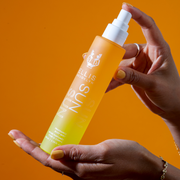 Model with yellow nail polish wearing silver ring and gold bracelets holding and spraying Sun Hair and Body Fragrance Mist 100ml without cap in both hands, on orange background.