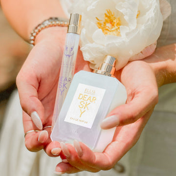 Modèle avec du vernis à ongles nu, portant des bagues en argent, des bracelets en or et en argent, et une robe blanche, tenant Dear Sky 10ml Eau De Parfum Spray de Voyage, Dear Sky 50ml Eau De Parfum avec le bouchon retiré, et une fleur blanche dans les deux mains.