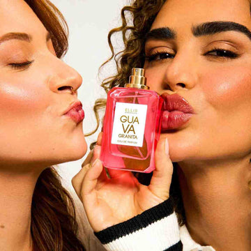 Model with brown curly hair and nude nail polish wearing black and white long sleeve sweater holds Guava Granit 50ml Eau De parfum with cap removed near lips and next to lips of model with red hair and gold nose ring wearing white sweater.