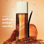 Miss Amber perfume mist 90mL bottle with a white cap, surrounded by amber liquid, saffron threads, vanilla bean, and stone chips on a warm peach background. Text on the left reads 