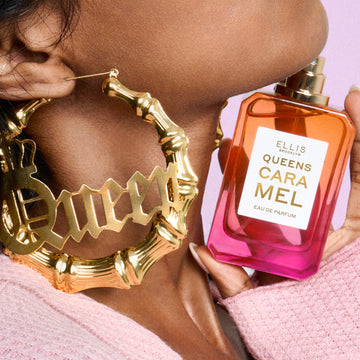 Gros plan d'une femme portant un pull rose et des boucles d'oreilles créoles en or où est écrit « Queen », tenant le parfum QUEENS CARAMEL Eau de Parfum 50 ml sans le bouchon sous son menton sur un fond rose