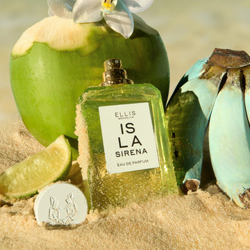 ISLA SIRENA Eau de Parfum 50ml without cap with sand on the bottle sits in sand next to it's cap, a slice of lime, a bunch of blue bananas, and a green coconut with a white flower on it
