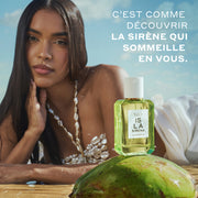 Un mannequin aux cheveux bruns portant un haut coquillage blanc et une jupe blanche est allongé sur le ventre dans le sable humide, la main sur le menton, derrière l'ISLA SIRENA Eau de Parfum 50 ml posée sur un coco vert avec un texte blanc qui dit « C'est comme découvrir la sirène qui sommeille en vous », le tout sur un fond de ciel bleu et de nuages blancs.