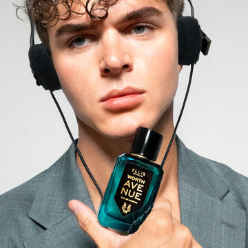 Model with brown hair wearing gray jacket and headphones holding Worth Avenue 50ml Eau De Parfum to chin, on white background.