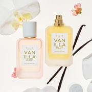 Bouteilles de 50 mL d'Eau de Parfum Vanille Riz et Vanille Sel entourées d'orchidées blanches, de fleurs roses et de gousses de vanille sur un fond blanc. Vanille Riz est présentée dans une bouteille rose pâle avec un bouchon blanc, et Vanille Sel dans une bouteille jaune doré avec un vaporisateur doré.