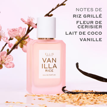 Eau de parfum VANILLA RICE (50 ml), présentée aux côtés de riz grillé, de gousses de vanille et de fleurs de cerisier (sakura) roses sur un fond rose, avec un texte gris indiquant : « Notes de riz grillé, fleur de cerisier, lait de coco, vanille ».