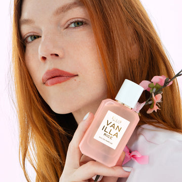 Une femme aux cheveux roux vêtue d'une chemise rose tient une eau de parfum VANILLA RICE (50 ml) et des fleurs de cerisier roses contre son épaule, sur un fond rose.