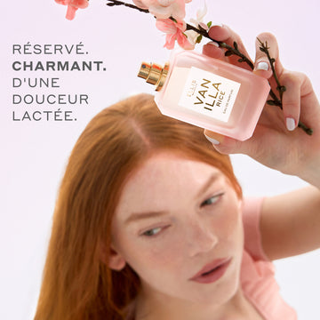 Une femme aux cheveux roux vêtue d'une chemise rose tient l'eau de parfum VANILLA RICE (50 ml, sans bouchon) ainsi que des fleurs de cerisier roses au-dessus de sa tête, sur un fond rose portant le texte gris : « Reserve. Charmant. D'une. Douceur. Lactee. »