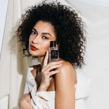 Model with black curly hair, nude nail polish, red lipstick, wrapped in white sheet against white fabric background holding Myth 50ml Eau De Parfum on shoulder.