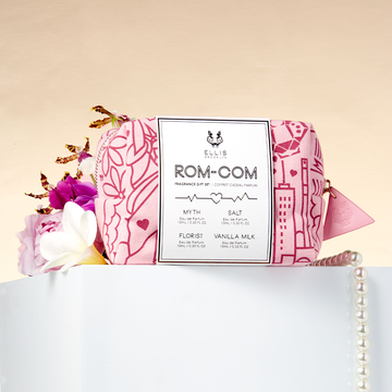 Sac cadeau parfum Rom-Com à motifs rouges et roses sur une table blanche avec un collier de perles, une fleur rose, une fleur violette, une fleur blanche et une fleur jaune, sur un fond orange clair.