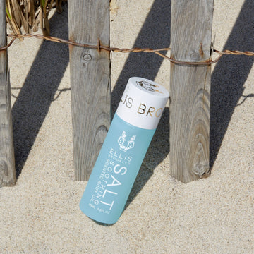 Salt 95ml Soothing Scented Body Oil in sand leaning against wooden and metal fence with beach grass in background.